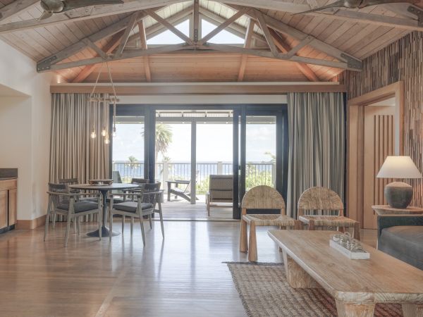 A spacious, rustic living area with wooden beams, dining table, sofa, and coffee table. Large windows open to a view and natural light.