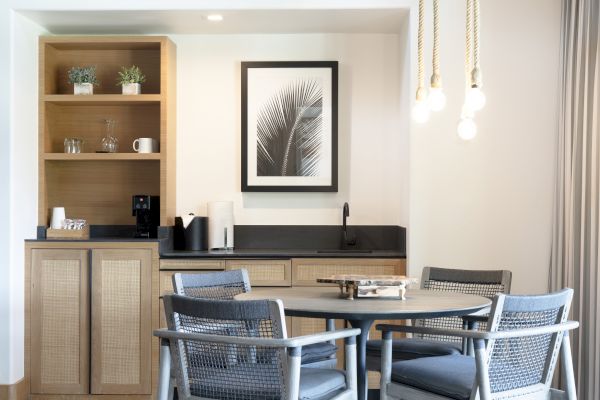 A modern dining area with a round table, chairs, a small kitchenette, and framed leaf art on the wall. Hanging lights add warmth.