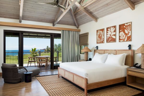 A cozy bedroom with a large bed, wooden ceiling, and ocean view through sliding doors. The room has a seating area and decorative wall art.