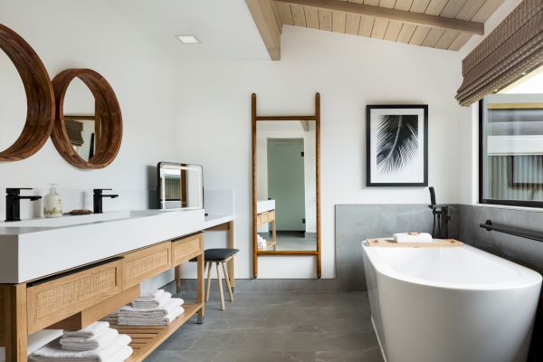 A modern bathroom features a freestanding tub, a vanity with two sinks, wooden accents, a large mirror, a stool, and a window with blinds.