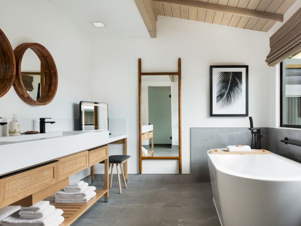 A modern bathroom features a freestanding tub, a vanity with two sinks, wooden accents, a large mirror, a stool, and a window with blinds.