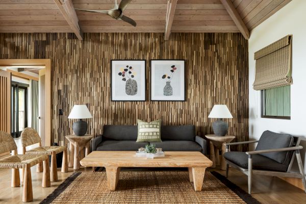 A cozy living room with a wooden accent wall, two framed artworks, a dark sofa, wooden tables, chairs, and neutral decor.