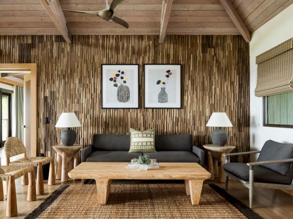 A cozy living room with a wooden accent wall, two framed artworks, a dark sofa, wooden tables, chairs, and neutral decor.