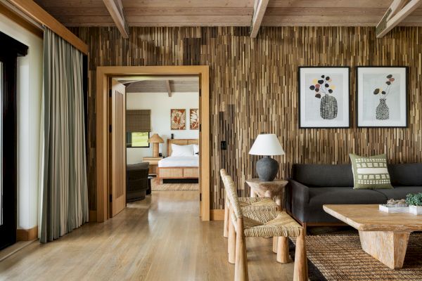 This image shows a cozy living area with wooden accents, a sofa, wooden tables and chairs, and a doorway leading to a bedroom with a visible bed.