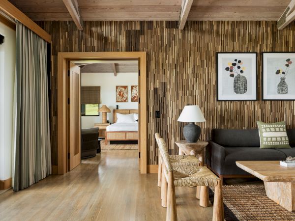 This image shows a cozy living area with wooden accents, a sofa, wooden tables and chairs, and a doorway leading to a bedroom with a visible bed.