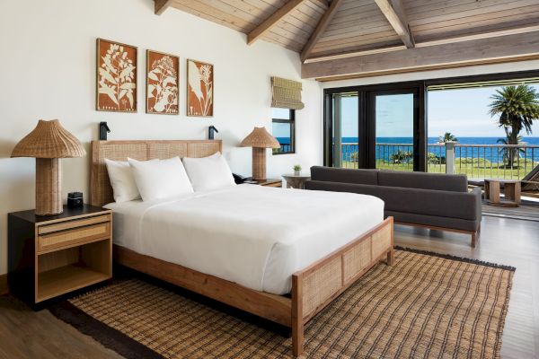 A cozy bedroom with a wooden bed, two side tables, lamps, framed art, a sofa, a large window overlooking the ocean, and a woven rug on the floor.