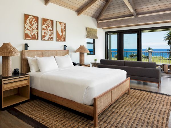A cozy bedroom with a wooden bed, two side tables, lamps, framed art, a sofa, a large window overlooking the ocean, and a woven rug on the floor.