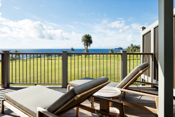 The image shows two lounge chairs on a wooden deck overlooking a grassy field with a view of the ocean and some palm trees in the background.