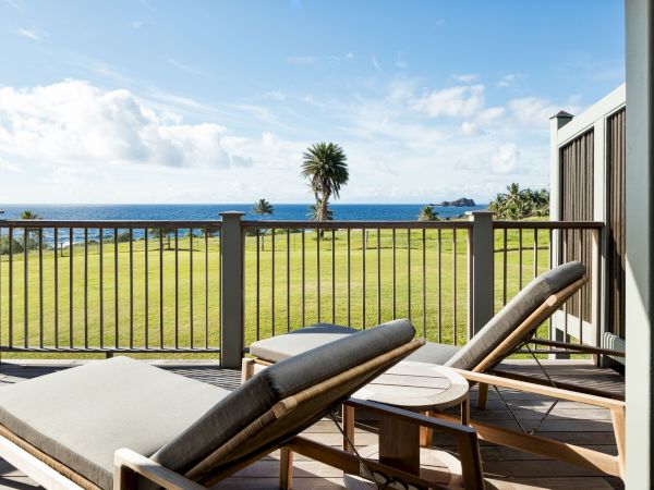 The image shows two lounge chairs on a wooden deck overlooking a grassy field with a view of the ocean and some palm trees in the background.