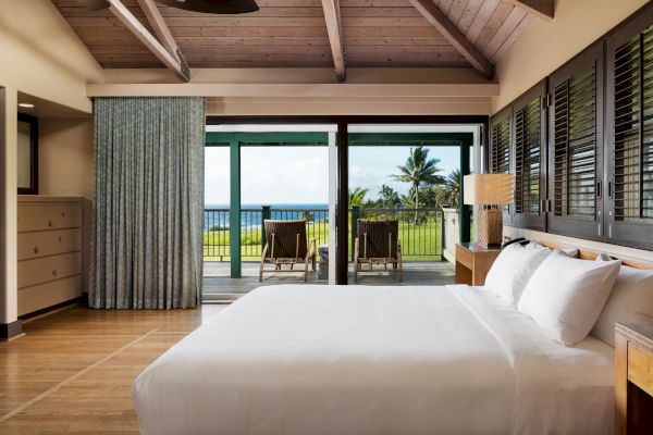 A bedroom with a bed, dresser, and sliding doors opening to a balcony with a view of the ocean and palm trees.