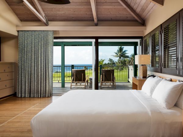 A bedroom with a bed, dresser, and sliding doors opening to a balcony with a view of the ocean and palm trees.