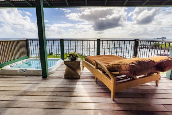 A deck with an ocean view, featuring a wicker lounge chair, a hot tub, and a small table under a partially covered roof.