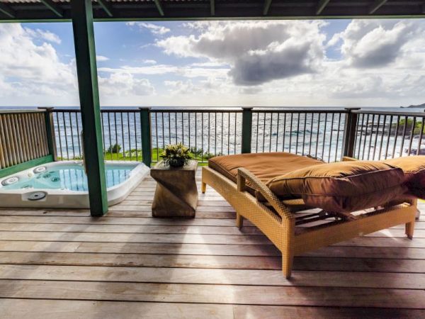 A deck with an ocean view, featuring a wicker lounge chair, a hot tub, and a small table under a partially covered roof.