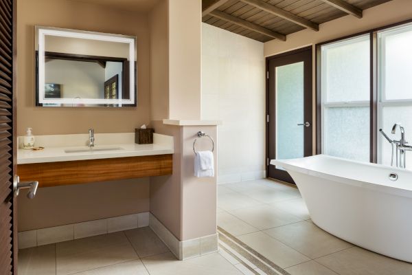 The image shows a modern bathroom with a freestanding bathtub, a sink with a mirror above it, a towel on a holder, and large windows.