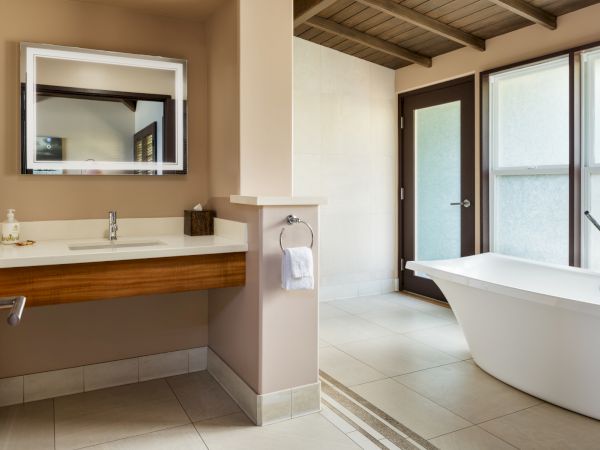The image shows a modern bathroom with a freestanding bathtub, a sink with a mirror above it, a towel on a holder, and large windows.