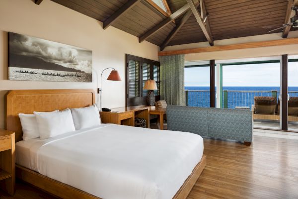 A cozy bedroom with a white bed, wooden furniture, and an ocean-view balcony, featuring large windows and a high wooden ceiling, is shown.