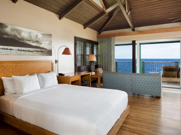 A cozy bedroom with a white bed, wooden furniture, and an ocean-view balcony, featuring large windows and a high wooden ceiling, is shown.