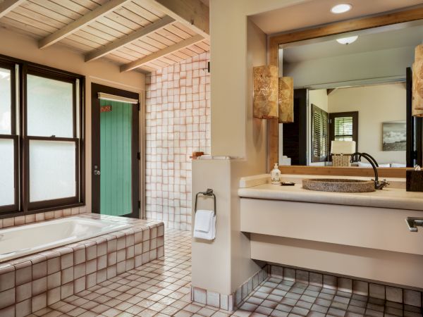 The image shows a well-designed bathroom with a bathtub, a large mirror above a sink, wooden ceiling beams, and a mix of tiles in earthy tones.