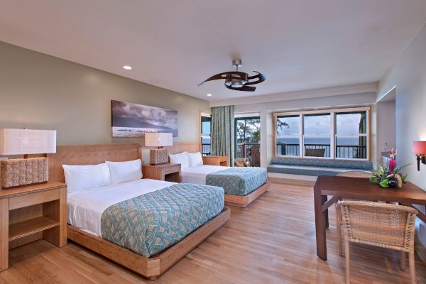 A spacious hotel room with two beds, a seating area, a desk, and a view of the ocean through large windows. The decor is modern and coastal.