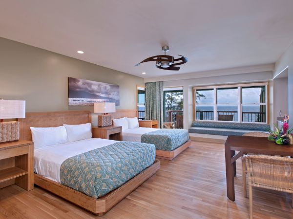 A spacious hotel room with two beds, a seating area, a desk, and a view of the ocean through large windows. The decor is modern and coastal.