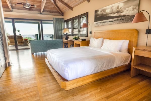 A bright bedroom with wooden floors, a comfy bed, a desk, lamps, and a balcony with a seating area and ocean view.