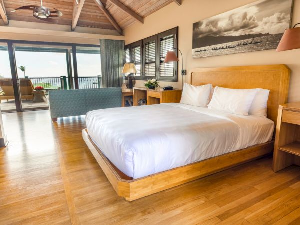 A bright bedroom with wooden floors, a comfy bed, a desk, lamps, and a balcony with a seating area and ocean view.