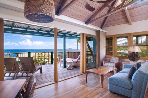 This image shows a cozy living room with wooden furniture and a sliding door opening to a balcony overlooking a beautiful ocean view.