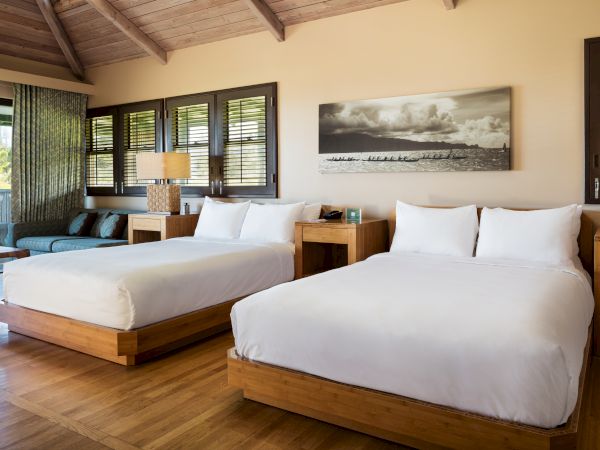 The image shows a well-lit hotel room with two beds, a wooden floor, a seating area, and a wall-mounted landscape picture.