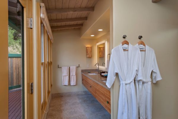 A bathroom with wooden ceiling, bathrobes on hooks, towels on a rack, mirrors, and sinks, and a door leading outside to a deck area.