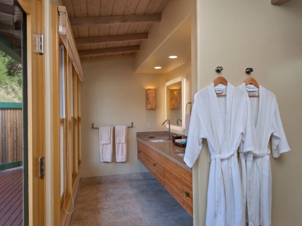 A bathroom with wooden ceiling, bathrobes on hooks, towels on a rack, mirrors, and sinks, and a door leading outside to a deck area.