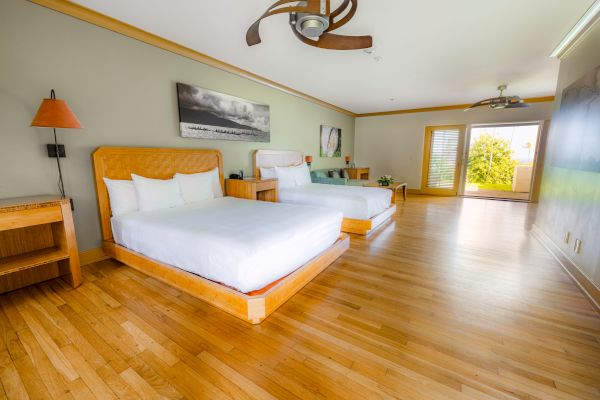 A spacious, modern hotel room features two large beds, wooden flooring, a ceiling fan, and a glass door opening to a patio with garden views.