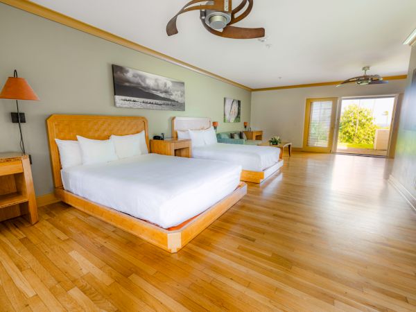 A spacious, modern hotel room features two large beds, wooden flooring, a ceiling fan, and a glass door opening to a patio with garden views.