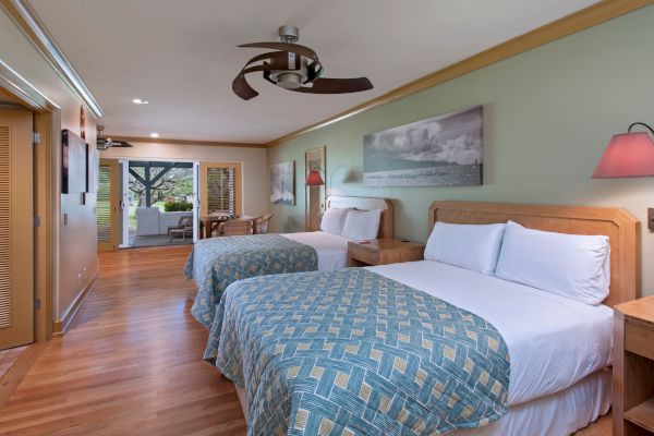 The image shows a hotel room with two beds, wooden furniture, modern decor, and a view leading out to a balcony or patio area with outdoor seating.