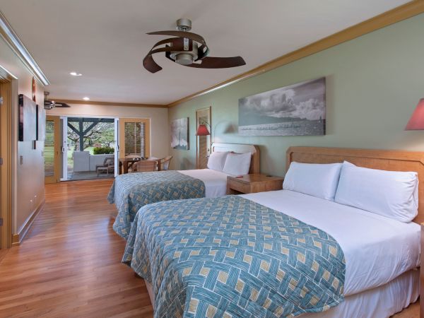 The image shows a hotel room with two beds, wooden furniture, modern decor, and a view leading out to a balcony or patio area with outdoor seating.