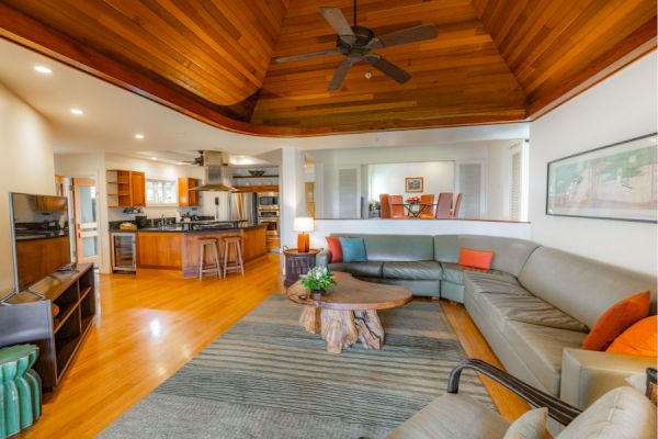 A spacious living room with a large sectional sofa, a rustic coffee table, an open kitchen, wooden ceiling, and modern furnishings.