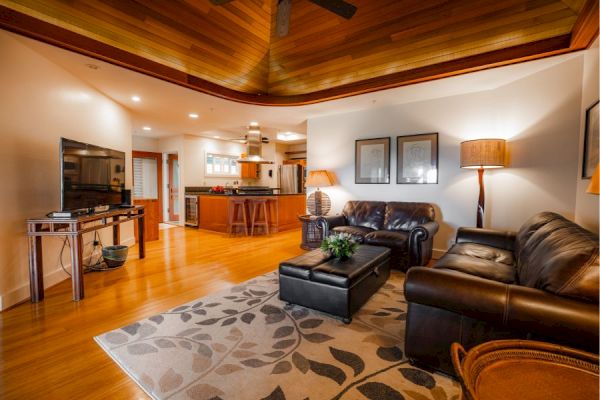 A cozy living room with leather couches, a TV, and a kitchen in the background. The wooden ceiling and warm lighting create a comfortable ambiance.