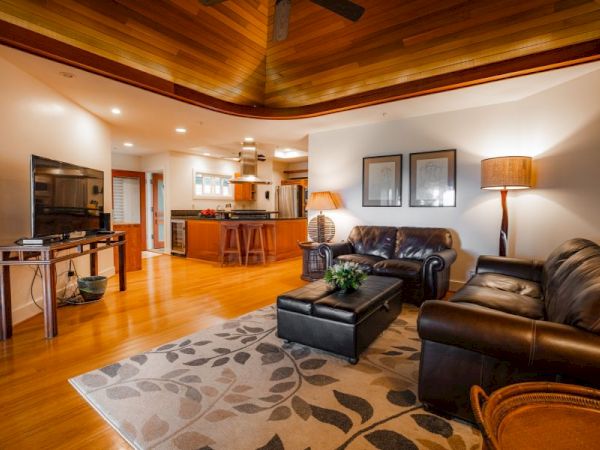 A cozy living room with leather couches, a TV, and a kitchen in the background. The wooden ceiling and warm lighting create a comfortable ambiance.