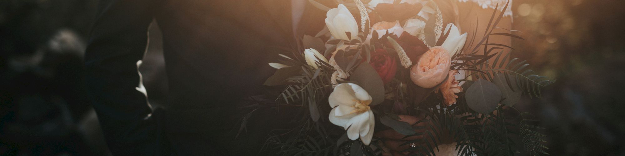 A couple stands together in wedding attire, with the bride holding a vibrant bouquet, surrounded by soft sunlight.