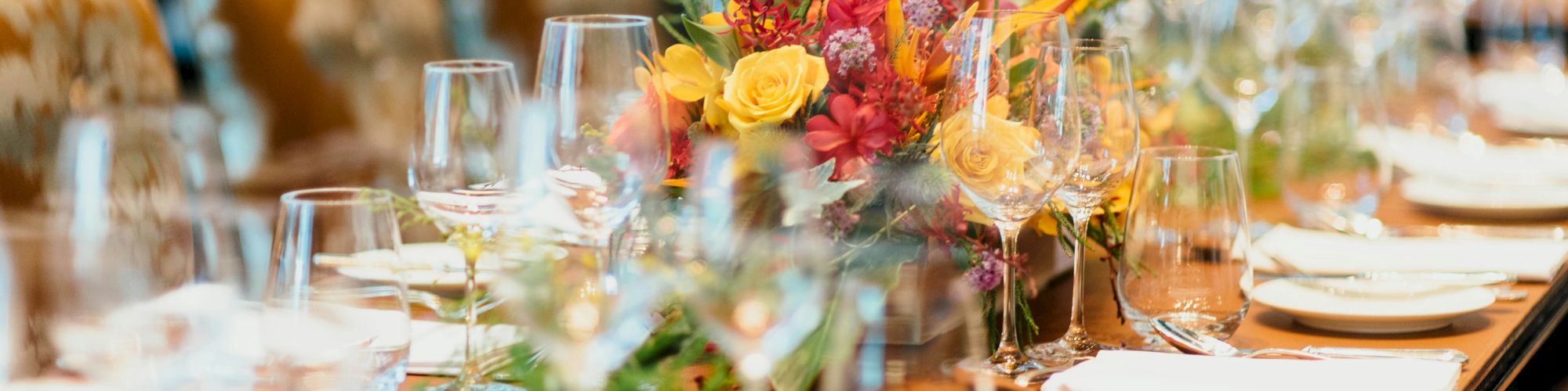 A long table is elegantly set with glassware, plates, and a vibrant floral centerpiece, ready for a formal dining event, ending the sentence.