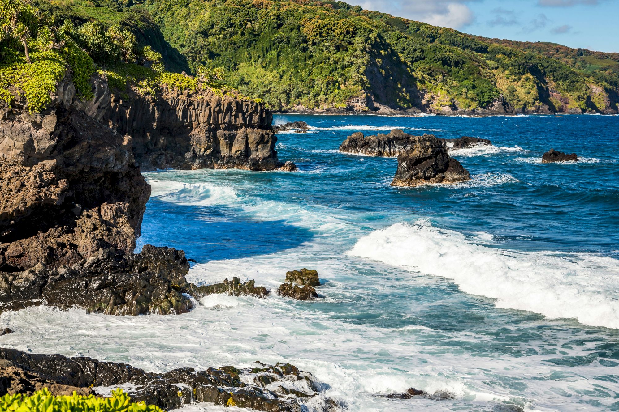 The image shows a rocky coastline with waves crashing against the rocks and lush green hills in the background under a blue sky.