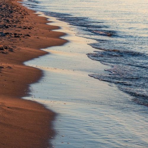 A sandy beach with gentle waves lapping at the shoreline, reflecting the soft light of either sunrise or sunset on the calm water, is depicted.