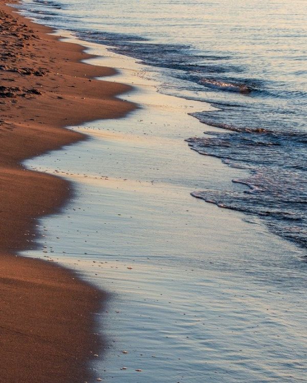 A sandy beach with gentle waves lapping at the shoreline, reflecting the soft light of either sunrise or sunset on the calm water, is depicted.