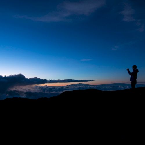 A silhouette of a person standing on a hill capturing the stunning twilight sky with scattered clouds and a subtle horizon in the background.