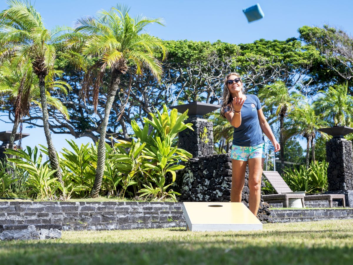 A person is playing a bean bag toss game outdoors, surrounded by lush greenery and palm trees.