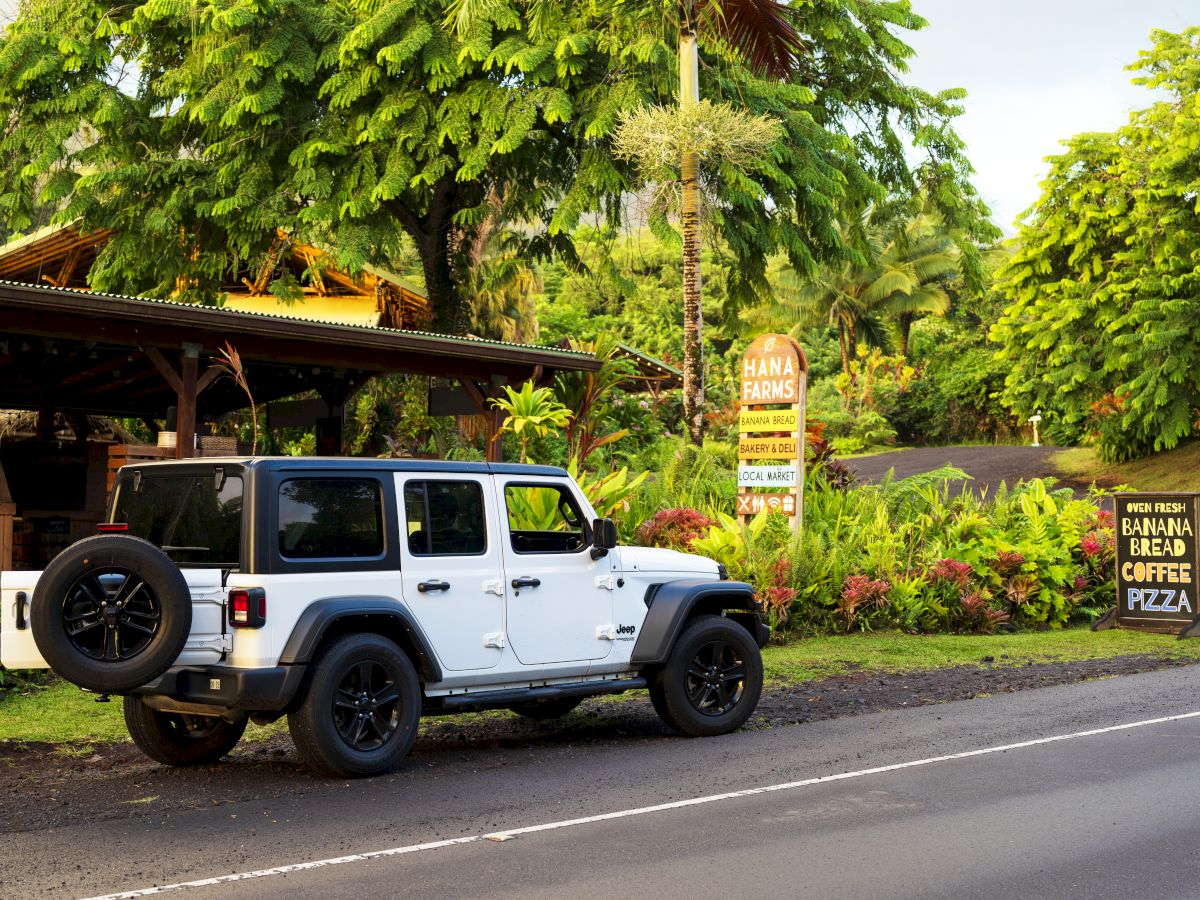 A white SUV is parked on a road beside a lush, green area with a café sign advertising banana bread, coffee, and pizza.