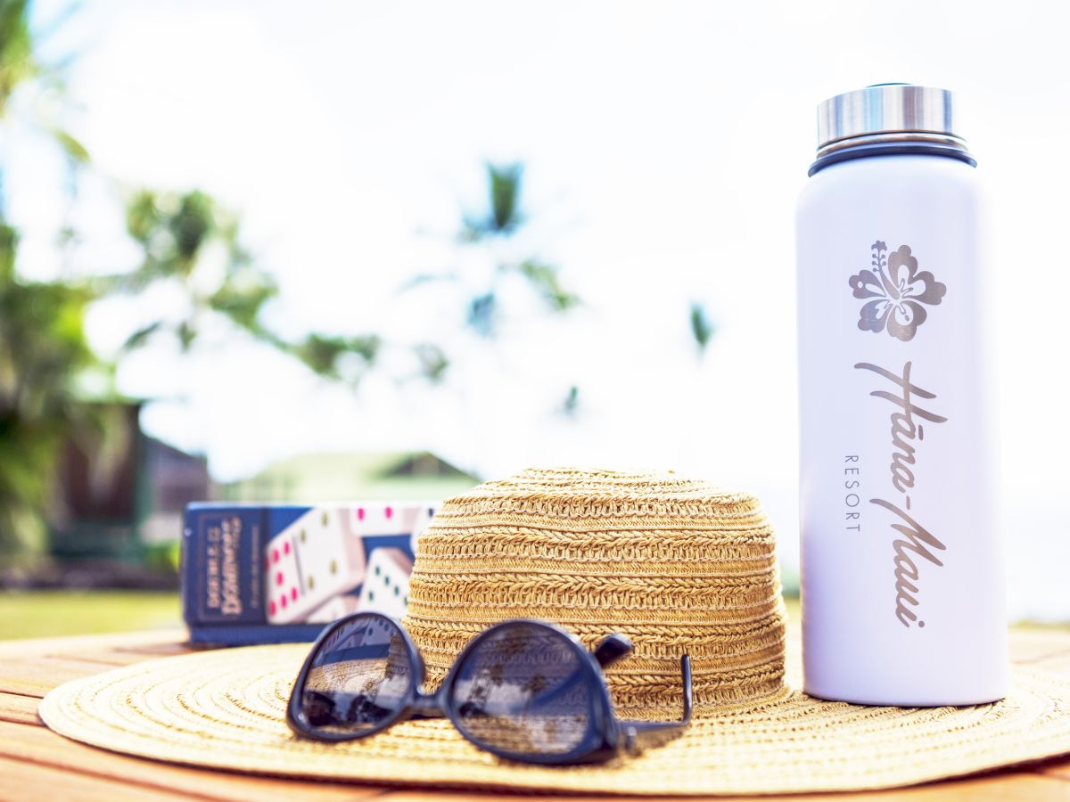 Sunglasses, a straw hat, a water bottle, and playing cards on a wooden table outdoors with a blurred background.