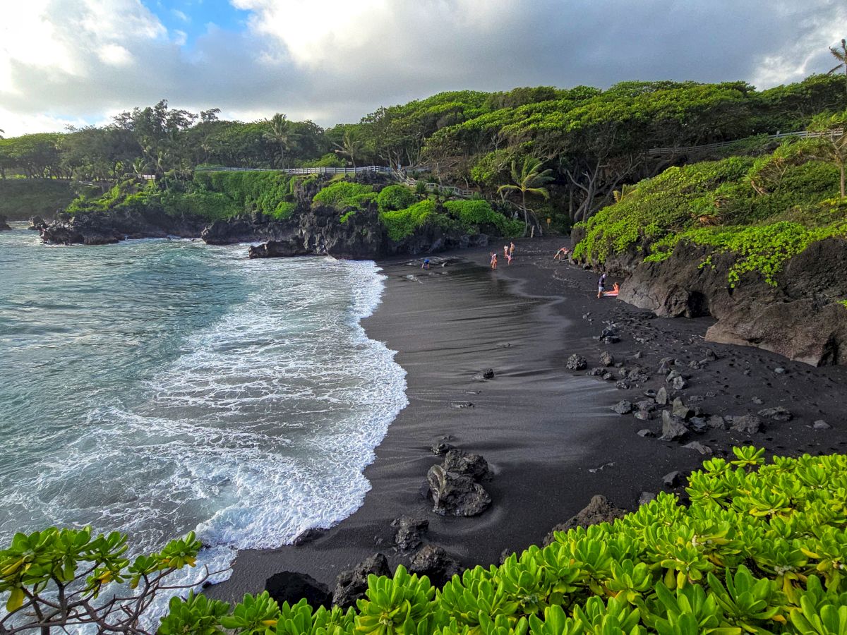 A black sand beach with lush greenery and waves crashing against the shore under a partly cloudy sky.