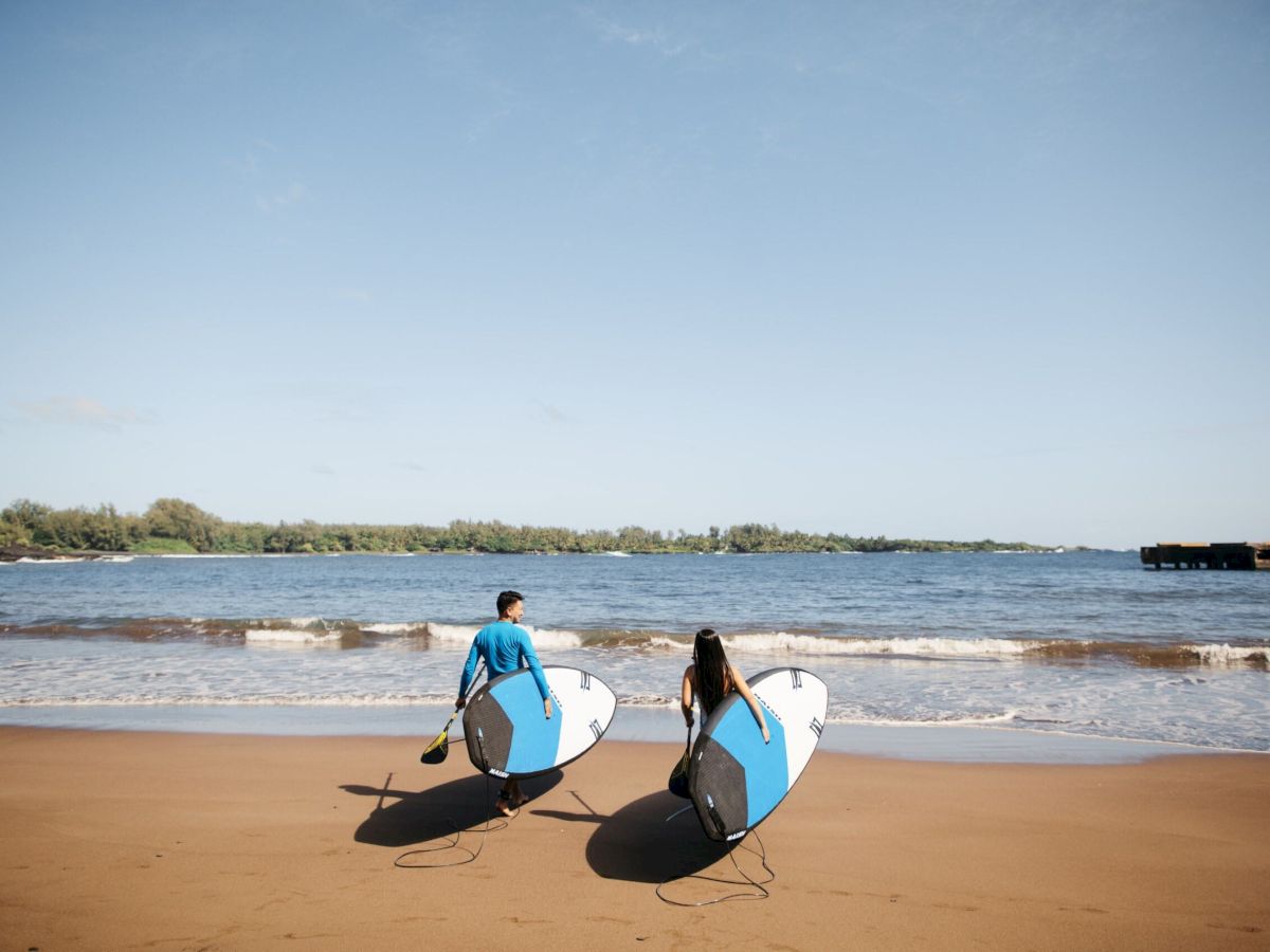 Two people are walking toward the ocean on a sandy beach, each carrying a surfboard and paddle, with a clear sky and distant shoreline in the background.