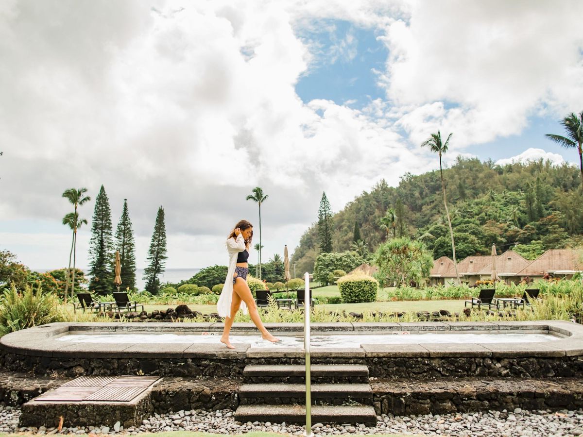 A person walking in a stone-edged pool area surrounded by greenery and mountains under a partly cloudy sky.