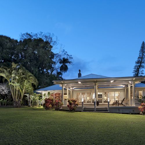 A charming, single-story house with a spacious porch is set against a backdrop of lush greenery and towering trees, illuminated at dusk.